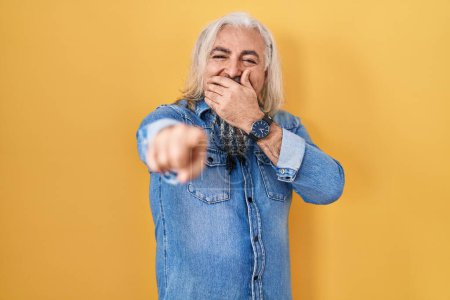 Photo for Middle age man with grey hair standing over yellow background laughing at you, pointing finger to the camera with hand over mouth, shame expression - Royalty Free Image