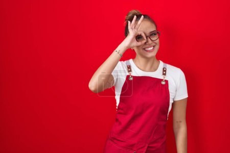 Téléchargez les photos : Jeune femme hispanique portant tablier de serveuse sur fond rouge faisant un geste correct avec la main souriante, les yeux regardant à travers les doigts avec un visage heureux. - en image libre de droit