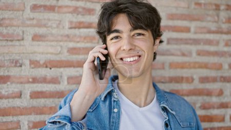 Téléchargez les photos : Jeune homme hispanique souriant confiant parler sur le smartphone sur fond de briques isolées - en image libre de droit