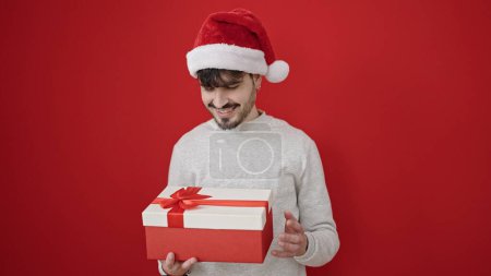 Téléchargez les photos : Jeune homme hispanique portant un chapeau de Noël déballer cadeau sur fond rouge isolé - en image libre de droit