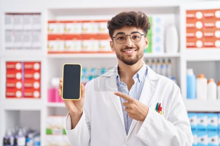 Téléchargez les photos : Arabe homme avec barbe travaillant à la pharmacie pharmacie montrant écran smartphone souriant heureux pointant avec la main et le doigt - en image libre de droit