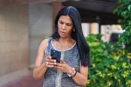 Téléchargez les photos : Moyen Age femme hispanique en utilisant un smartphone avec une expression sérieuse dans la rue - en image libre de droit
