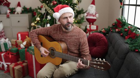 Photo for Young bald man playing classical guitar sitting on sofa by christmas tree at home - Royalty Free Image