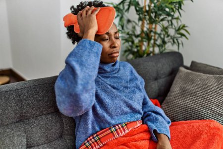 Photo for African american woman holding hot water bag on head sitting on sofa at home - Royalty Free Image