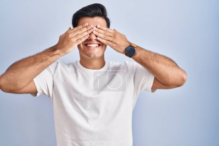 Photo for Hispanic man standing over blue background covering eyes with hands smiling cheerful and funny. blind concept. - Royalty Free Image