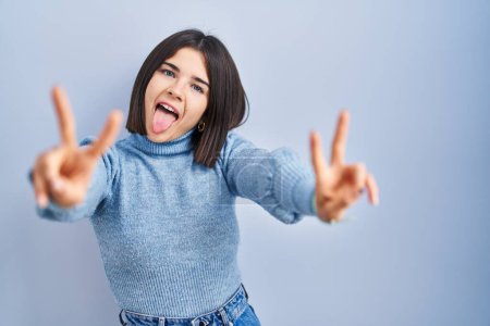 Photo for Young hispanic woman standing over blue background smiling with tongue out showing fingers of both hands doing victory sign. number two. - Royalty Free Image