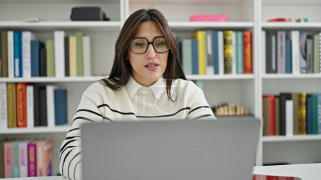 Foto de Joven hermosa estudiante hispana usando laptop con expresión seria en la universidad de la biblioteca - Imagen libre de derechos