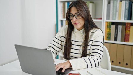 Téléchargez les photos : Jeune belle étudiante hispanique utilisant un ordinateur portable avec une expression sérieuse à l'université de la bibliothèque - en image libre de droit