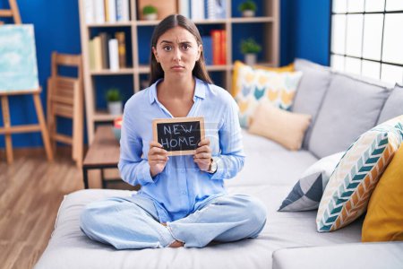 Photo for Young hispanic woman holding blackboard with new home text sitting on the sofa skeptic and nervous, frowning upset because of problem. negative person. - Royalty Free Image