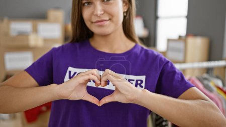 Photo for Radiant young hispanic volunteer woman confidently flashing a heart gesture, standing happily in a bustling charity center filled with donations - Royalty Free Image