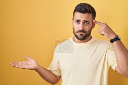 Photo for Handsome hispanic man standing over yellow background confused and annoyed with open palm showing copy space and pointing finger to forehead. think about it. - Royalty Free Image