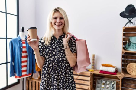 Foto de Joven mujer rubia sonriendo confiada sosteniendo bolsas de compras y café en la tienda de ropa - Imagen libre de derechos