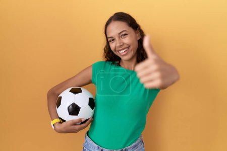 Photo for Young hispanic woman holding ball approving doing positive gesture with hand, thumbs up smiling and happy for success. winner gesture. - Royalty Free Image