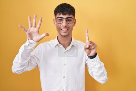 Téléchargez les photos : Jeune homme hispanique debout sur fond jaune montrant et pointant vers le haut avec les doigts numéro six tout en souriant confiant et heureux. - en image libre de droit