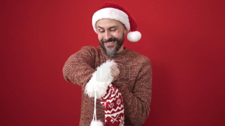 Photo for Young bald man wearing christmas hat looking inside of sock over isolated red background - Royalty Free Image