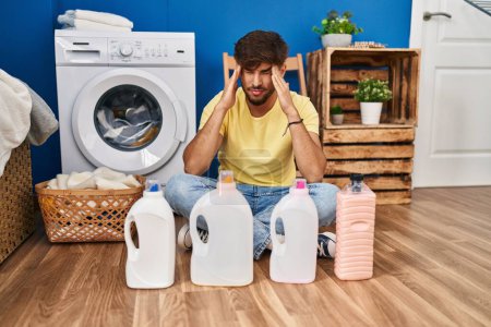 Photo for Arab man with beard doing laundry sitting on the floor with detergent bottle with hand on head, headache because stress. suffering migraine. - Royalty Free Image