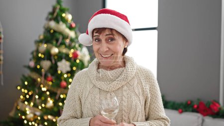 Photo for Mature hispanic woman speaking to the camera drinking wine standing by christmas tree at home - Royalty Free Image