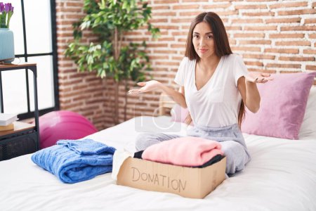 Photo for Young hispanic woman putting used clothes into donations box sitting on the bed clueless and confused expression with arms and hands raised. doubt concept. - Royalty Free Image