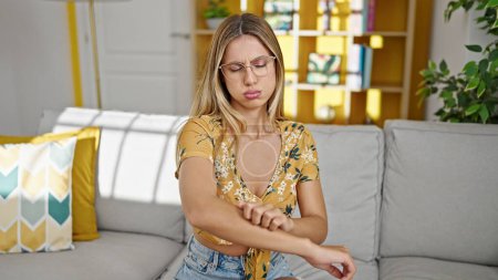 Foto de Mujer rubia joven sentada en el sofá rascarse el brazo para picar en casa - Imagen libre de derechos