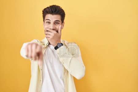 Téléchargez les photos : Jeune homme hispanique debout sur fond jaune se moquant de vous, pointant du doigt la caméra avec la main sur la bouche, expression de honte - en image libre de droit