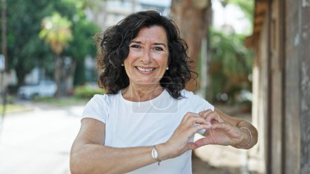 Foto de Mujer hispana de mediana edad sonriendo confiada haciendo gesto de corazón con las manos en el parque - Imagen libre de derechos