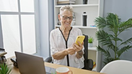 Téléchargez les photos : Femme d'affaires aux cheveux gris souriante tapant un message joyeux sur son smartphone au bureau - en image libre de droit