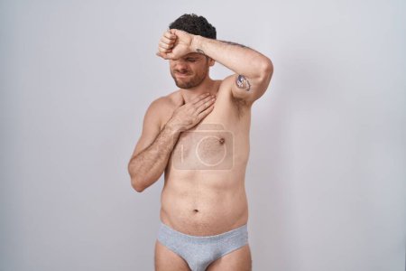 Téléchargez les photos : Jeune homme hispanique debout torse nu portant des sous-vêtements touchant le front pour la maladie et la fièvre, la grippe et le rhume, le virus malade - en image libre de droit