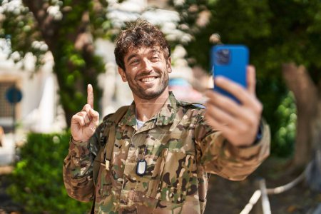 Téléchargez les photos : Jeune homme hispanique portant un uniforme de camouflage de l'armée faisant appel vidéo souriant avec une idée ou une question pointant du doigt avec un visage heureux, numéro un - en image libre de droit
