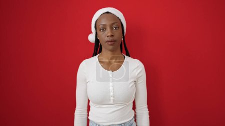 Téléchargez les photos : Femme afro-américaine portant un chapeau de Noël debout avec une expression sérieuse sur fond rouge isolé - en image libre de droit