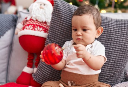 Photo for Adorable hispanic baby holding maraca and ball sitting on sofa by christmas tree at home - Royalty Free Image