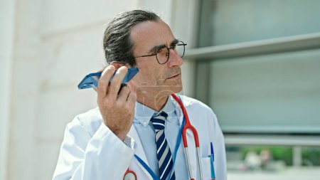 Photo for Middle age man doctor listening to voice message at hospital - Royalty Free Image
