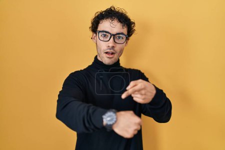 Téléchargez les photos : Homme hispanique debout sur fond jaune à la hâte pointant pour regarder le temps, l'impatience, bouleversé et en colère pour retard de délai - en image libre de droit
