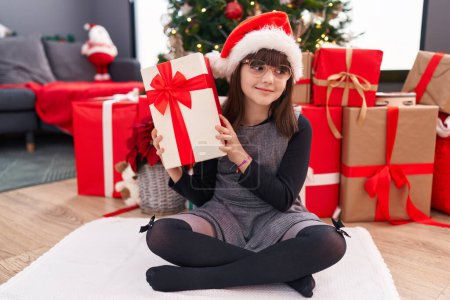 Foto de Adorable chica hispana sosteniendo regalo de Navidad sentada en el suelo en casa - Imagen libre de derechos