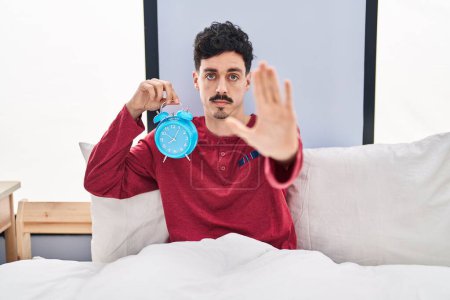 Photo for Hispanic man holding alarm clock in the bed with open hand doing stop sign with serious and confident expression, defense gesture - Royalty Free Image