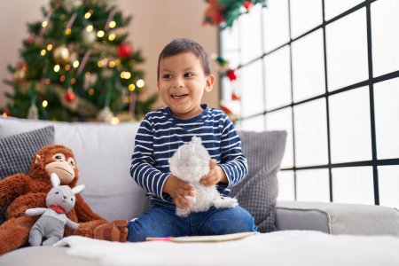 Foto de Adorable niño hispano jugando con juguetes sentados en el sofá junto al árbol de Navidad en casa - Imagen libre de derechos