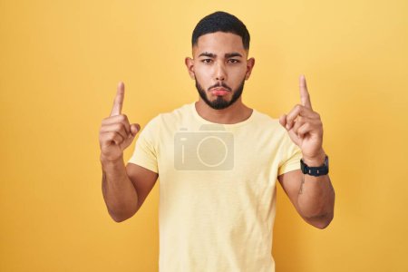 Téléchargez les photos : Jeune homme hispanique debout sur fond jaune pointant vers le haut regardant triste et contrarié, indiquant la direction avec les doigts, malheureux et déprimé. - en image libre de droit