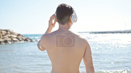 Téléchargez les photos : Jeune homme hispanique touriste écoutant de la musique debout à l'envers au bord de la mer - en image libre de droit