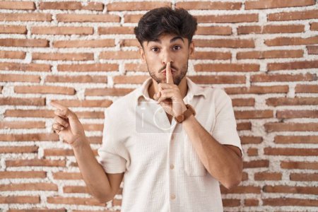 Photo for Arab man with beard standing over bricks wall background asking to be quiet with finger on lips pointing with hand to the side. silence and secret concept. - Royalty Free Image