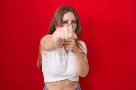Photo for Young caucasian woman standing over red background punching fist to fight, aggressive and angry attack, threat and violence - Royalty Free Image