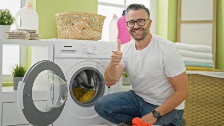 Téléchargez les photos : Homme aux cheveux gris lavant des vêtements faisant un geste de pouce souriant à la buanderie - en image libre de droit