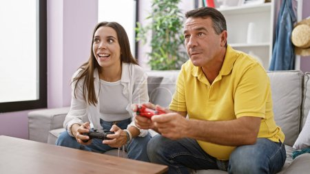 Téléchargez les photos : Père hispanique et fille aiment coller le temps de jouer à un jeu vidéo à la maison, assis sur le canapé du salon, sourit de joie sur leurs visages, comme ils utilisent avec confiance leurs commandes de manette de jeu. - en image libre de droit
