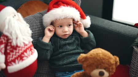 Téléchargez les photos : Adorable bambin blond assis sur le canapé portant un chapeau de Noël à la maison - en image libre de droit