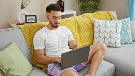 Photo for Young arab man using laptop sitting on sofa at home - Royalty Free Image