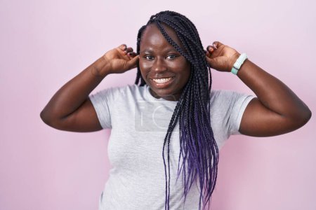 Photo for Young african woman standing over pink background smiling pulling ears with fingers, funny gesture. audition problem - Royalty Free Image