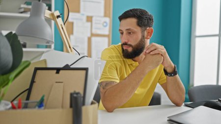 Téléchargez les photos : Jeune homme hispanique étant viré assis sur le bureau regardant paquet d'effets personnels au bureau - en image libre de droit
