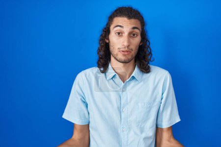 Photo for Young hispanic man standing over blue background clueless and confused with open arms, no idea concept. - Royalty Free Image