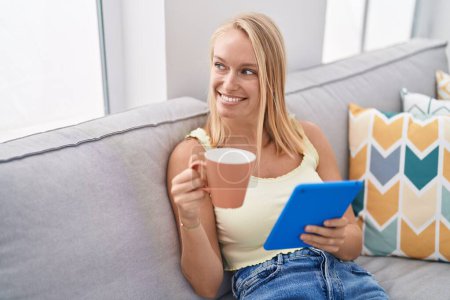 Foto de Mujer rubia joven bebiendo café y usando touchpad en casa - Imagen libre de derechos