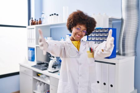Téléchargez les photos : Jeune femme afro-américaine travaillant au laboratoire scientifique regardant la caméra sourire à bras ouverts pour se faire câliner. expression joyeuse embrassant le bonheur. - en image libre de droit