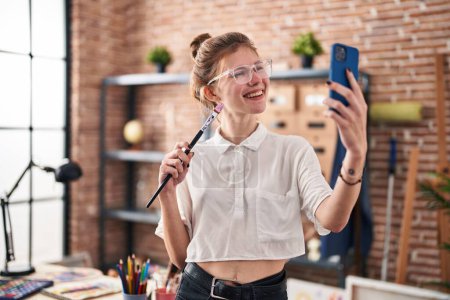 Téléchargez les photos : Jeune femme blonde artiste tenant pinceau faire sefie par smartphone au studio d'art - en image libre de droit