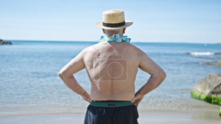 Téléchargez les photos : Senior homme aux cheveux gris touriste en maillot de bain et chapeau d'été debout vers l'arrière au bord de la mer - en image libre de droit